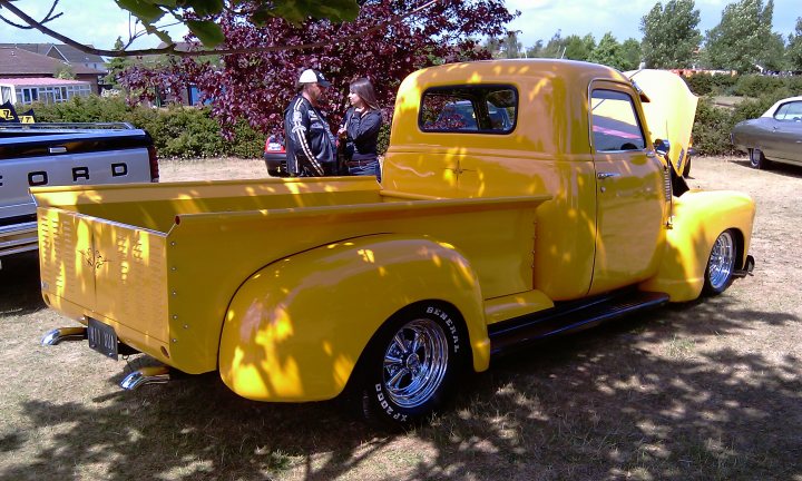 Cars On The Green - Photo Thread - Page 1 - East Anglia - PistonHeads - The image features a large, old-style passenger truck painted in bright yellow. One older couple and another individual are standing next to the truck, admiring or posing with the vintage vehicle. The truck has an open cargo bed at the back and is parked in an open area adjacent to some trees. Another older car can be seen in the background, also parked. It seems to be a sunny day as there is ample sunlight illuminating the scene.