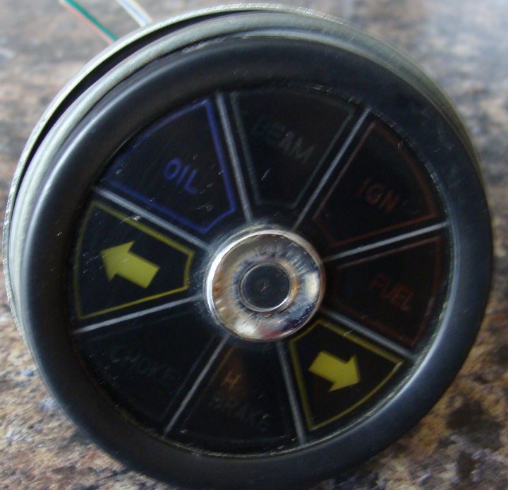 Instrument Cluster Pistonheads - The image shows a round object with a six-sided arrangement of panels. Each panel has a different symbol and color. Starting from the top left and moving clockwise, the symbols on the panels include an oil drop, a fuel droplet, an ignition symbol, a fire symbol, a fire symbol, and an upward arrow. The arrows and text urge the insertion of fresh fuel into a vehicle, indicating exhaust and other types of fuel. The object is black with colored and textured insets that provide indicators for different automotive components.