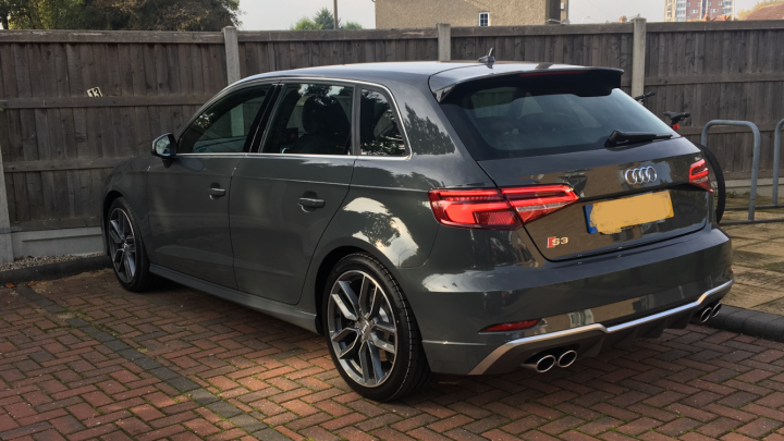 The Audi S3 lease thread - Page 8 - Audi, VW, Seat & Skoda - PistonHeads - The image shows a gray Audi hatchback parked in a lot. The car is positioned diagonally across the photo, viewed from a slightly elevated angle that includes the front and back doors. The design of the car is modern, and it features a distinctive rear wing for increased aerodynamics. On the license plate area, there is a visible logo and registration plate. In the background, there are fences and a building partially obscured, indicating the location is likely a public parking area or a street-side parking spot.