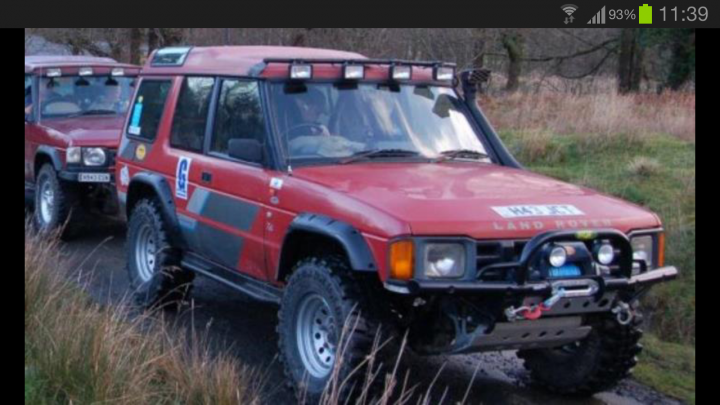 STOLEN Land Rover Discovery from Kingswood - Page 1 - South West - PistonHeads - The image features a red Land Rover Defender 90 (shown on the side of the vehicle) with its lights on, parked on the side of a gravel road. The car has a four-light alarm system on the front and several other cars can be seen in the background, suggesting that this is a convoy or a line of vehicles. The rear of the vehicle is facing the camera, providing a view of its windshield, windows, and side mirrors. There's a person in the background, likely nearby, but they are not the focus of the image.