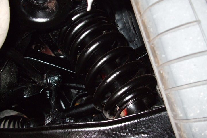 Pistonheads Underseal Waxoyl - This image captures an inside view of a vehicle's engine bay. The primary focus is on part of the vehicle's suspension spring. The spring is wound and compacted, suggesting the location could be from beneath the vehicle's hood. The background is dark, indicating the interior of the vehicle's engine compartment. The surrounding area is cluttered with various mechanical parts and components, including wiring and pipes, which further indicate the inner workings of a vehicle's engine. The photo seems to have been taken with a close-up lens, emphasizing the details of the mechanical parts.