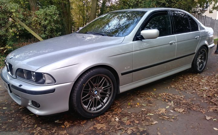 Show Me Your BMW!!!!!!!!! - Page 411 - BMW General - PistonHeads - The image shows a silver-colored BMW car parked outdoors. It appears to be a four-door sedan model with distinctive styling elements such as the kidney grille, large headlights, and multi-spoke alloy wheels. There is a visible black soft top roof that seems to be in good condition. The vehicle has a set of custom rims on the front wheels, which add a sporty touch to its appearance. The car's bodywork shows some modifications, possibly for aerodynamics or aesthetics, given the smooth and streamlined design. In the background, there is a leafy tree and what looks like a wooden fence, indicating that this photo was taken in a suburban or semi-rural environment.