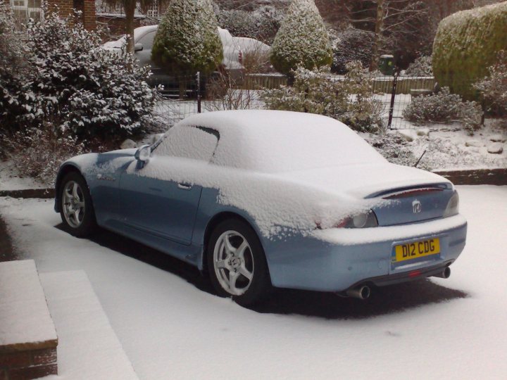 Light Pistonheads Touchpaper Nurburgring Stand Blue - The image captures a rear view of a light blue car beautifully covered in a thick blanket of snow. The car's sporty lines are accentuated by the snow, and its dark blue wheels are uniformly covered in snow. The wintery backdrop is a mix of snow-covered bushes and a smattering of bare trees, creating a serene and peaceful winter scene. The car's license plate is visible and reads "DIZ CDG", indicating its registration details.