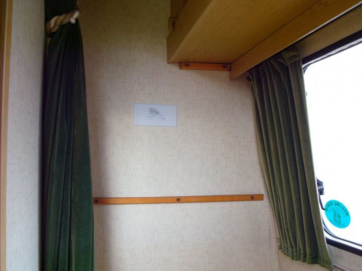 My caravan history - Page 1 - Tents, Caravans & Motorhomes - PistonHeads - The image shows an interior view of a small room with a curtain. On the wall, to the left of the curtain, there is a dark green fabric which hangs like a valance. Below this fabric, along the baseboard, is a long wooden bar or rail that matches the baseboard and the window trim. Adjacent to the rail, there is a white object attached to the wall; however, it's unclear what it is due to the resolution of the image. The window on the far right side of the photo features a bright sky view, and the curtain's fabric looks green with a darker shade on its right, closest to the window.