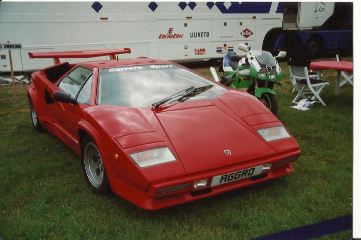 Countach  - Page 12 - Lamborghini Classics - PistonHeads - The image captures a classic red Porsche 911 GT1 with a large fixed wing, parked in a grassy field. The car is positioned next to a white tent, which has advertising fascia covering its side. There's a table with chairs nearby, suggesting an event or gathering. A green motorcycle is also visible in the background, adding to the impression of a gathering or exhibition at this location.