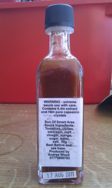 Hot Pistonheads Sauce - The image features a red pepper sauce that's shaped like a bottle. The sauce has a yellowish-brown color with a red pepper flake floating on top, indicating its spicy contents. On the label, there's a warning in black text which includes warnings about its extreme hotness and specific usage instructions. The label also includes the name "Son of Smart Ase" and mentions the ingredients and amount (weight or liquid) per bottle.