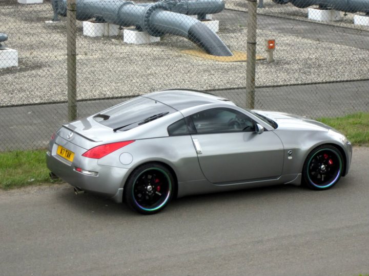 V12V contrasting roof color, opinions? - Page 1 - Aston Martin - PistonHeads - The image depicts a scene on a dusty road where a silver sedan is parked. The car is positioned on the driver's side of the road, facing away from the camera, suggesting a routine parking situation. The environment around the car includes a metal fence to the left side, a sidewalk, and a grassy area, indicating a well-maintained roadside. The presence of two pipes in the vicinity gives a hint of a possible construction or maintenance project nearby.