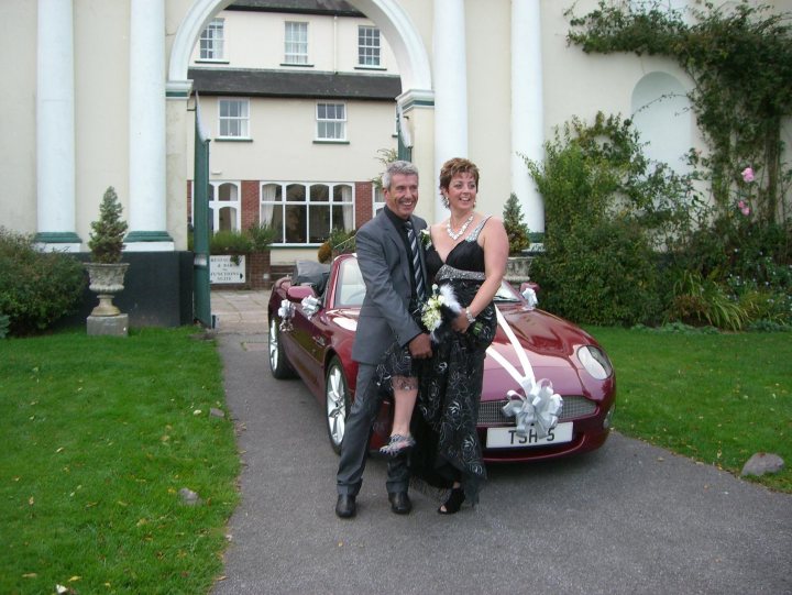 Pistonheads - The image features a joyous moment in front of what appears to be a church or a building with an archway and green shrubs. A man and a woman are standing on a pathway, posing for the photograph with the woman partially draped over the man. Both are smartly dressed in suits and evening gowns. The woman is holding a bouquet of flowers and there is a car with a ribbon bearing numbers, suggesting an upcoming event, possibly a wedding or a ceremony. The sky is overcast, providing soft lighting to the scene.