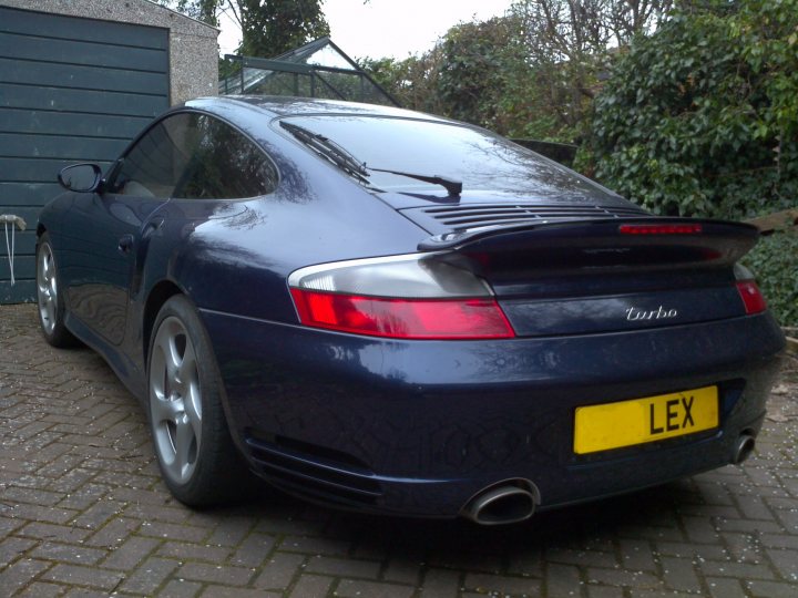 show off your porsche - Page 1 - Porsche General - PistonHeads - The image showcases a black Porsche parked on a brick driveway. The car is a sleek sports model, known by the logo "Porsche" on the trunk. Key details include a distinctive rear spoiler and a red taillight that adds to its striking appearance. A yellow license plate reading "LEX" can be seen, and the car is parked in front of a garage. The tail end of the vehicle is prominently displayed, highlighting its aerodynamic design.