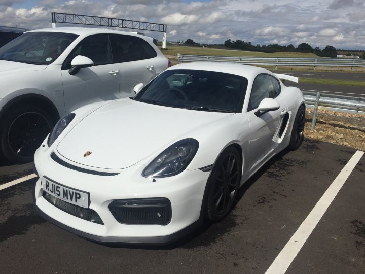 GT4 colours  - Page 52 - Boxster/Cayman - PistonHeads - The image shows a white Porsche sports car parked in a parking lot with white lines. The car's design is sleek and modern, with a distinct rear spoiler and alloy wheels. The Porsche is parked next to another white vehicle, which appears to be a station wagon or SUV. The background features a partially cloudy sky and what seems to be a runway or an open field, suggesting that the parking lot may be adjacent to an airport or an aviation facility.