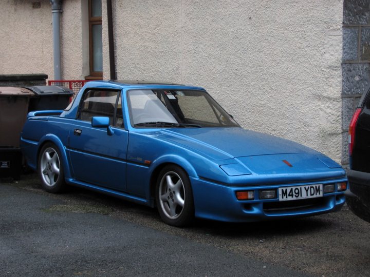 Pistonheads - The image showcases a vintage blue two-door sports car parked on the side of the road. The car, dating back to the 1980s, is characterized by its silver diamond grill and black rubber bumpers. It's positioned in front of a white stone residential building, reflecting the car's minor scratches and adding to its classic appearance. A trash can is also visible near the car, indicating a public or residential setting. The car and its surroundings suggest a quiet neighborhood scene.