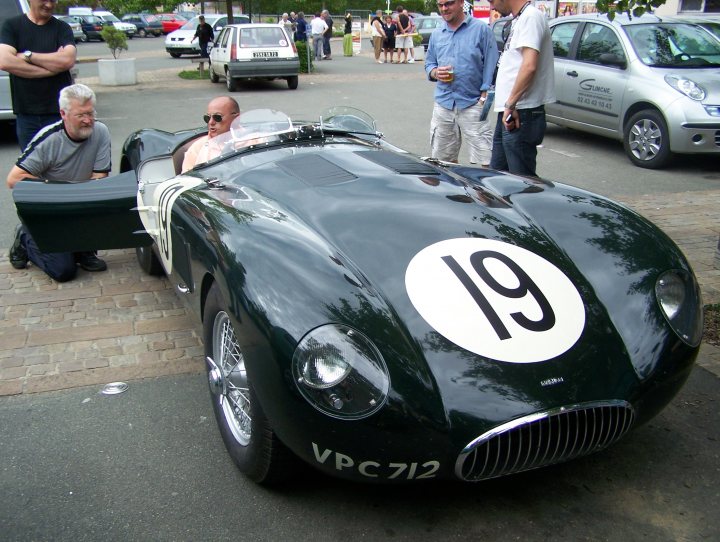 Jaguar Fans Pistonheads Strictly - This is an intriguing scene featuring a vintage car, likely an Alfa Romeo, given the distinctive round emblem on the hood with the number 19 on it. The car is parked and surrounded by a few individuals, dressed casually in t-shirts and jeans, who seem to be gathering around and marveling at the car. One person is even taking a photograph of the car. Other cars and a truck can be seen in the background, suggesting that this may be happening at some event or show. The setting appears to be an outdoor area with a brick surface, indicating it might be a public space like a park or a museum parking lot.