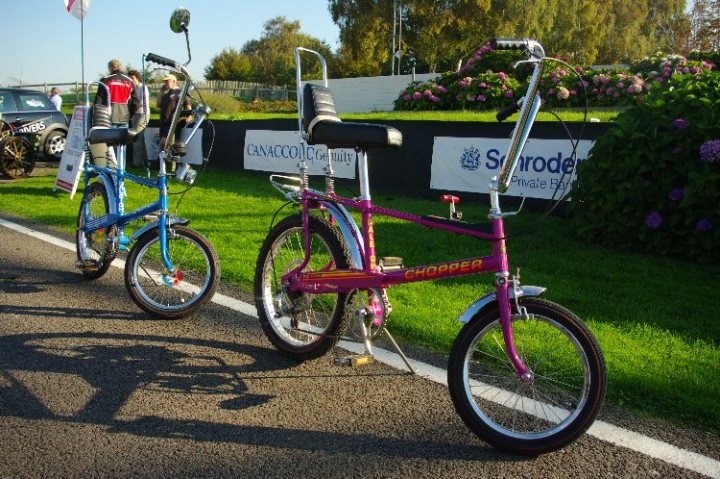 breakfast club sunday october 2 - Page 1 - Goodwood Events - PistonHeads - The image showcases a tranquil outdoor setting with two colorful bicycles parked on the side of a shaded street. The bicycles, one pink and the other blue, exhibit a classic style with visible curved fenders and frame comfortable for riding. In the backdrop, behind the bicycles, there's a lush green lawn and a fence adorned with a banner. The scene radiates a sense of leisure and calming atmosphere.