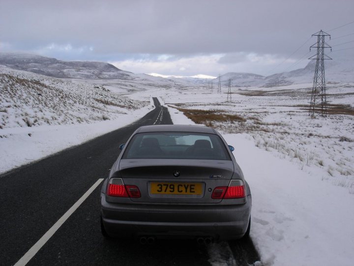 In the image, a gray BMW, bearing the license plate 379 CYE, is stopped on the side of a highway during winter. The car is positioned under a power line, suggesting it's at a junction or rest stop. The surrounding landscape shows more snow and that the photo was taken in early spring, given the presence of patches of snow and a rough, uneven road surface. The sky is cloudy, adding to the overall wintry atmosphere.