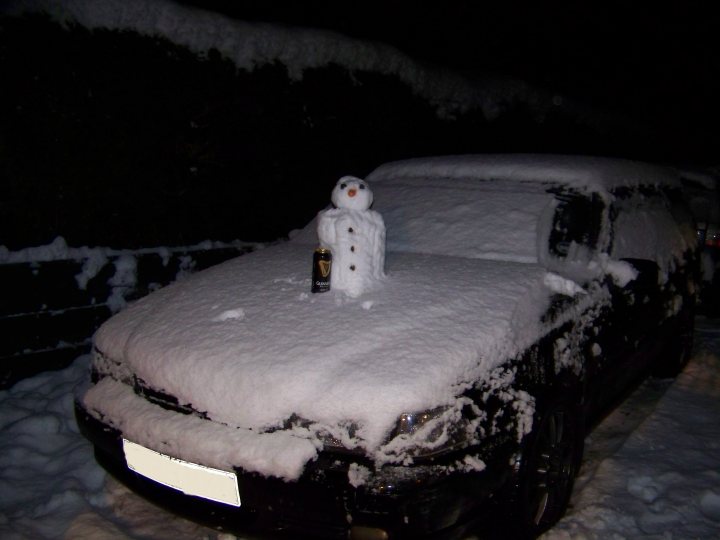 Pistonheads - The image presents a snowy scene, with a parked black car being the main subject. On top of the car, a snowman stands proudly, its smiley face visible above the blanket of snow. The car is parked on a driveway, which is mostly covered by a mound of snow. A bottle of beer, frozen in the snow, rests next to the car. The overall scene is quirky and humorous, with the beer bottle and snowman adding a touch of whimsy to the otherwise ordinary winter setting.