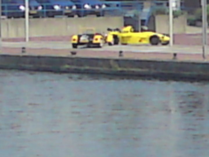 Interesting spot - The Hague - Page 1 - Netherlands - PistonHeads - The image captures a serene scene on a city river. A small boat is adrift, taking up a portion of the water body that appears calm despite the overcast sky. The river is enclosed by a levee running parallel to the water's edge, providing a boundary for the boat's journey.

In the background, closer to the other side of the river, a gold-colored racecar is parked. Its vibrant color contrasts with the muted tones of the overcast sky and the silhouetted outlines of trees. Despite the mention of a car accident, the image does not show any signs of distress or emergency. The overall scene suggests a quiet, possibly early morning or late evening, atmosphere.