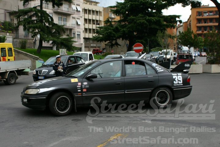 Banger Rallies - Page 1 - Events/Meetings/Travel - PistonHeads - This image captures a bustling city street, specifically in Paris. On the street, a black car with a distinct black and white number 39, possibly indicating a racing or sporting event number, is driving under a red and white no entry sign. Two people are standing on the sidewalk next to the black car. The atmosphere suggests a lively cityscape with various vehicles, including a truck in the background, parked along the street, and a building with an orange facade can be seen on one side of the street. The image conveys a sense of movement and life in the urban setting.