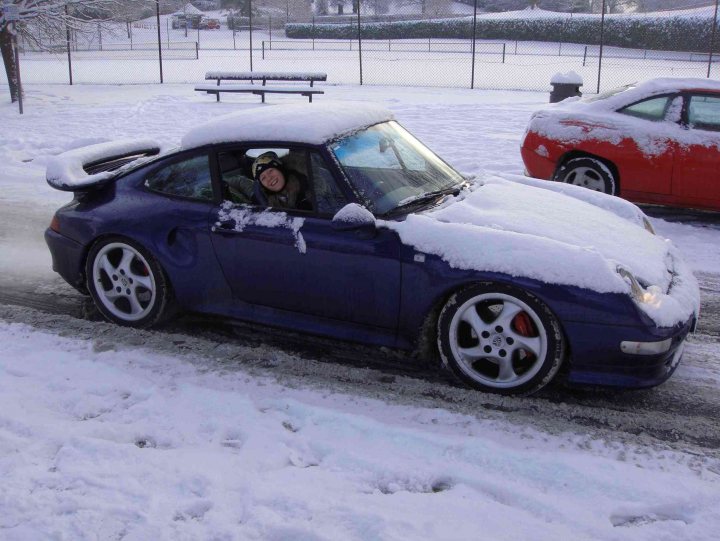 993 TURBO 'S'..... - Page 1 - Porsche General - PistonHeads - In the midst of a snowy field, a blue sports car stands out. The car's hood is dusted with snow, indicating a recent fall. A solitary figure can be seen through the car's window, suggesting the car is not abandoned but simply parked. Two other cars faintly visible in the background, along with a solitary bench, give context and hint at human activity. The image captures a moment of tranquility amidst the winter chill.