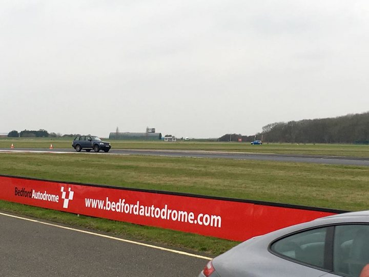 The Range Racer - Page 2 - Readers' Cars - PistonHeads - This image shows a view of a motor racing track. In the foreground, there's a sign that reads "Bedford Autodrome" along with its website: "www.bedfordautodrome.com". The background reveals a grassy infield and what appears to be a paved racing area with various cars on it, suggesting an active race or practice session. There is also a car parked close to the camera, and the sky overhead is overcast.