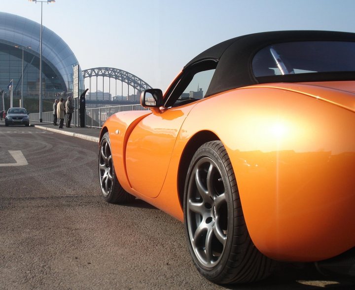 Random TVR pics - Page 1 - General TVR Stuff & Gossip - PistonHeads - This image captures a vibrant street scene. In the foreground, an orange sports car is parked by the sidewalk. The car's sleek design and shiny paint reflect the light, adding a dynamic element to the image. Further down the street, another car is following behind the sports car. The perspective of the photo is interesting, taken from the rear of the sports car as if viewer is standing behind it. In the background, a large building looms, possibly suggesting an urban setting. The sky above is a clear blue, suggesting a sunny or pleasant day.