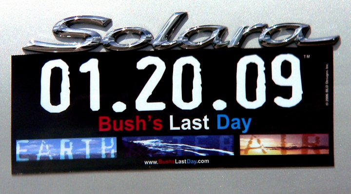 The image features a black bumper sticker with white text. The sticker displays the date "01/20/09" and reads "SOLARA Bush's Last Day". At the bottom of the sticker, there are images of Earth, ice, and water, and the website "www.lastday.com" is also mentioned. This sticker may be expressing a political message or commemorating a specific day in history. It's unusual to find a car with a sticker this particular, as it references a specific event in U.S. history.