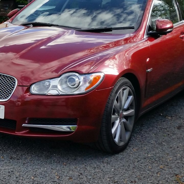 Buyer beware - approved used - Page 1 - Jaguar - PistonHeads - The image features a red sedan parked on a paved surface. The car has sleek lines and a modern design, with decorative silver accents along the side. It is equipped with chrome-finished wheels and aftermarket headlights. The sky in the background is filled with clouds, suggesting that it might be a cloudy day. The vehicle appears to be in good condition, with no visible damage or scratches.