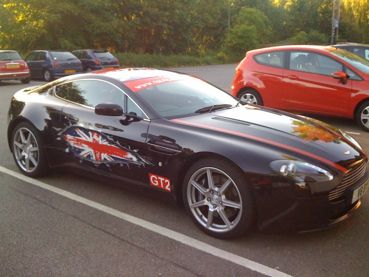 Stickered Pistonheads - The image features a striking black sports car, distinguished by its silver wheels and a red stripe running down the center. Parked in a parking lot with other cars present, this sleek vehicle is placed diagonally across the frame, facing towards the right. The car bears a distinctive British flag design on its side, hinting at its origin or ownership. The overall scene captures a casual setting with a hint of sophistication provided by the flag and the classic design of the sports car.