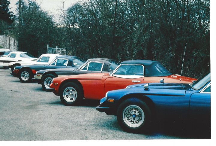 Early TVR Pictures - Page 32 - Classics - PistonHeads - The image shows a row of vintage cars parked closely together. From left to right, the first car is a bright white Bentley, followed by six black and red classic models with a prominent roadster design. The paint of the cars has a glossy finish, indicating good maintenance and care. The vehicles are lined up in a lot, suggesting they might be part of a collection or a special event. The setting appears to be a dedicated parking area for these cars, as there is no surrounding infrastructure or environment visible in the image.