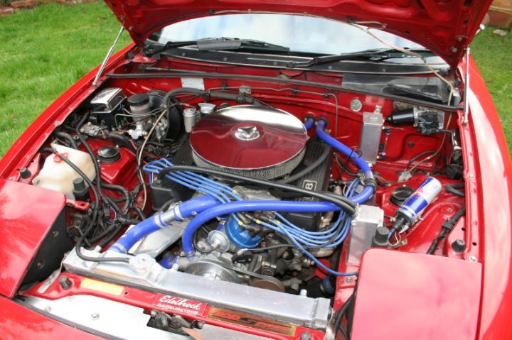 What do you drive? How much attention? - Page 29 - General Gassing - PistonHeads - The image provides a close-up view of the underside of a red sports car's engine compartment. The engine housing showcases a mix of mechanical parts, hoses, and tubes, with vibrant blue fluid connecting various components. A large heat shield, characterized by its circular design, covers a significant part of the engine, likely for thermal protection. A striking feature is the silver emblem, located near the center of the engine, which stands out against the maroon hue. The scene captures the intricate complexity of a high-performance vehicle's vital components, representing not just a car, but a testament to precision engineering.