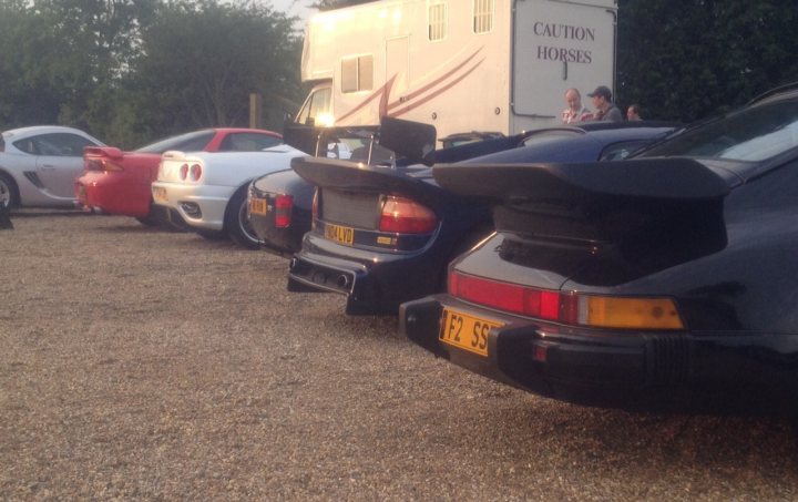 A bunch of cars that are parked in a lot - Pistonheads - The image showcases a gathering of people with an array of sports cars parked on what appears to be a rocky, outdoor terrain. Four sports cars, each with a distinct design, are prominently featured in the foreground. Each car has unique characteristics such as body color, model, and additional modifications. Additionally, there is a fifteenth-century living history event truck in the background, indicating the possible context of the vehicles' gathering. The attendees seem to be either inspecting the cars or engaging in conversations around them.