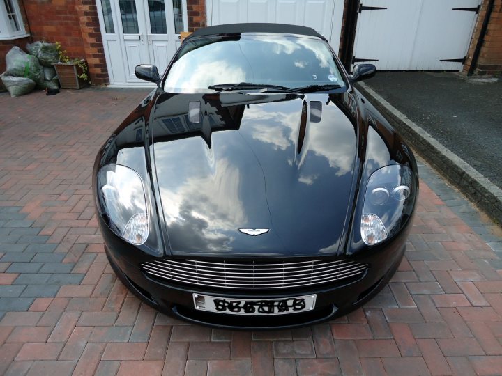 Pistonheads Detailed - The image features a sleek black sports car, most likely a convertible, parked in a driveway. The reflection of a partly cloudy sky in the car's glossy finish is noticeable, adding to the shiny appearance of the vehicle. The car is a Jaguar with a silver grille, as indicated by the logo on the front. In the background, there's a brick driveway leading to a white garage door, and a few planters with greenery are visible in the corner. There is also a license plate that reads "DIRECT GR8VX."