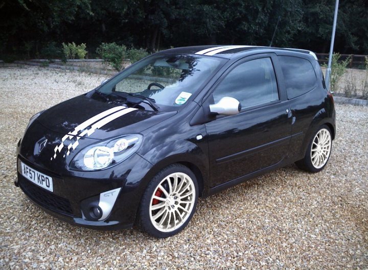 Twingo Suspension Pistonheads - The image shows a small, three-door black hatchback car parked on a gravel surface. The car features a striking diagonal stripe running across its hood, along with silver rims. The car's body has a glossy finish, indicating it might be well-maintained or recently washed. The glare of sunlight on the car suggests it may be a sunny day. The car is facing towards the left side of the photo.