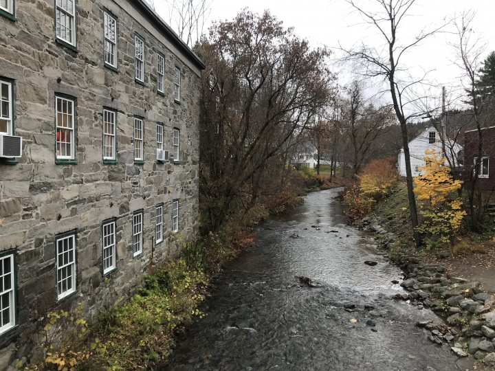 A red fire hydrant sitting in the middle of a forest - The image depicts a serene scene of a small town setting. A meandering creek flows through the middle of the frame, its waters reflecting the surrounding greenery and buildings. On either side of the creek are stone buildings with visible windows, one of which has an American flag flying on it. The architecture suggests that these buildings might be old or made in a style that harks back to a bygone era. The overall color palette is muted, with greens and browns dominating the scene, lending a calm and peaceful atmosphere to the image.