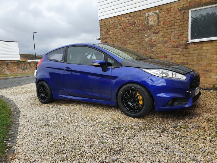 Fiesta ST MK7.5 Track car  - Page 31 - Readers' Cars - PistonHeads UK - The image shows a blue Ford car parked on a gravel surface, outside of what appears to be a residential house. The vehicle has modifications such as a body kit and aftermarket wheels, which gives it a sporty appearance. The car is positioned at an angle to the camera, showcasing its side profile. In the background, there is a building with a red brick facade. The sky is overcast with gray clouds, suggesting a cloudy or possibly rainy day. There are no visible texts or distinctive marks that would indicate the brand of the car or any other specific information.