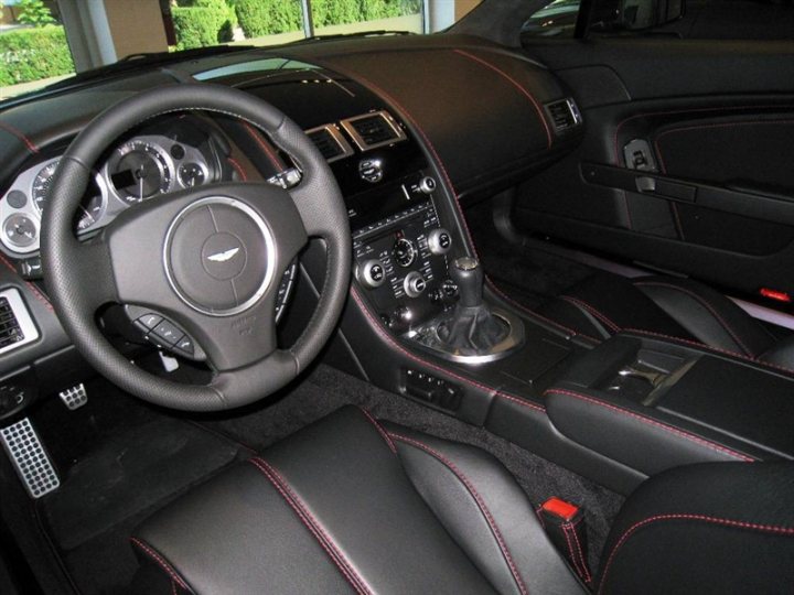 Did it! - Page 1 - Aston Martin - PistonHeads - The image shows the interior of a high-end luxury vehicle. It is a three-quarters perspective from the side, showing various controls, displays, and upholstery designs. The dashboard features a mix of analog and digital gauges and controls, with a consistent color palette of red, black, and silver. The car is equipped with leather upholstery with red stitching, which contrasts with the shiny black plastic and metallic accents. The steering wheel is large with multiple controls, indicating advanced driving and information features. This image represents a very stylish and sophisticated vehicle.