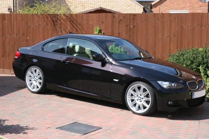 Deep black/maroon metallic, what to use seal/wax a new car? - Page 1 - Bodywork & Detailing - PistonHeads - The image depicts a black BMW car parked on the side of a brick-laid driveway. The vehicle is positioned towards the right of the frame, with the front facing towards the camera. The BMW logo is clearly visible on the front of the car. There is a wooden fence behind the car, and green foliage is visible beyond the fence, suggesting a residential setting. The overall scene is quiet and still, with no people or animals present in the image.