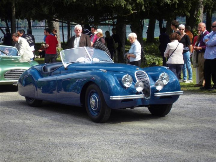 Pistonheads Deste Concoursporn Villa - This image captures a classic car show on a sunny day. A vintage convertible, painted in a striking shade of blue, is prominently displayed in the foreground. Its open-top design and shiny finish suggest it is well cared for. Surrounding the car is a crowd of people, dressed in casual attire, who appear to be enjoying the occasion. Other attendees are gathered further back, absorbed in the discussion of the displayed cars. The show is held outdoors, with the greenery and the sunshine adding to the lively atmosphere of the event.