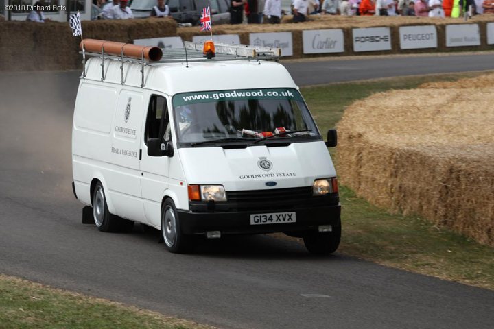 Pistonheads Drivers Van - The image captures a dynamic scene on a road likely designated for car shows, as indicated by the hay bails lining the track route. In the center of the image, a silver van is the main focus. It has a distinctive design element: an umbrella-like contraption on its roof, under which there's a pile of hay. This vehicle is the first among a line of others, suggesting that it might be the leader in some sort of race or procession.