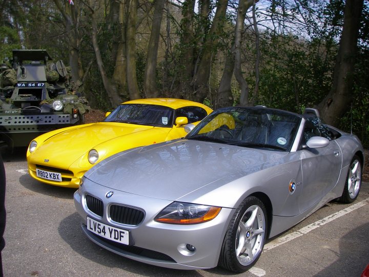 Pistonheads - The image features a gathering of vehicles in what appears to be a serene outdoor setting with trees in the background. On one side of the frame is a bright yellow car, which looks like a sports car, characterized by its sleek design and a distinctive black stripe. This vehicle is facing the camera, giving a clear view of its front and side. On the other side is a grey convertible with a distinguishable circular emblem on the side, representing a well-known luxury car brand. The grey car is also facing the camera, but it's slightly angled with the front hood slanted forward. There is a single person partially visible at the bottom left of the image, likely taking the photo. There is no visible text or distinctive markings on the vehicles that indicate distinctive brands or model types. There are no visible people interacting with the vehicles in the scene, and the background doesn't contain any individuals or buildings that would provide additional context about the location or event. The overall atmosphere is calm and seems unrelated to activities involving the cars. The parking arrangement of the cars suggests that this might be a casual gathering or an outdoor event where vehicle enthusiasts would display their trees.