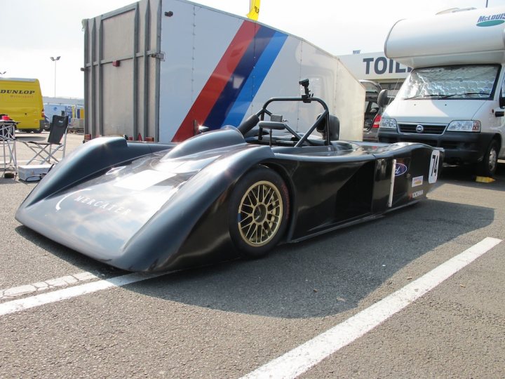 Pictures of your racecar thread - Page 1 - UK Club Motorsport - PistonHeads - The image features a drag racing car, which appears to be ready for a competition. The car is sleek and aerodynamically designed, with a wide, extended front end. Multiple sponsor logos are visible on the side of the car and on the rear wing. In the background, there is a large truck and another smaller vehicle, typical of the pits in a racing event. The sky is overcast, suggesting an overcast day, and there are no people immediately visible in the scene.