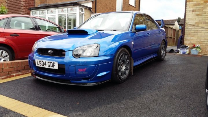 RE: Subaru Impreza WRX STI Type UK: PH Fleet - Page 3 - General Gassing - PistonHeads - The image shows a blue car parked on the side of a street. It appears to be a modified or customized vehicle, as indicated by its lowered stance and aftermarket parts such as the exhaust system and wheel modifications. The car is positioned next to a paved area with a curb, and there are two other vehicles visible in the background, one on each side. The surroundings suggest it's an urban setting with a residential neighborhood. There are no people visible in the image.