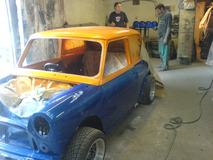 my little mini - Page 1 - Readers' Cars - PistonHeads - In a warm, rustic workshop, a vintage blue car with an orange roof is undergoing a meticulous restoration process. The car, featuring a distinctive chopped and channeled custom job, is in the process of having its top paint job applied. Two men are present in the scene, one is engaged in the act of painting the car's roof, while the other stands by, observing the process. A few other restoration projects are scattered in the background, hinting at a creative and skilled work environment.