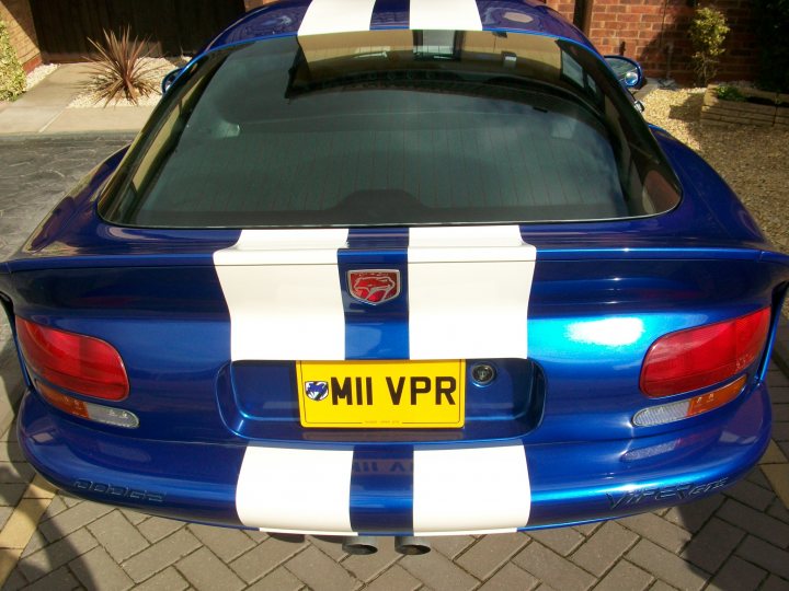 Pistonheads - The image shows the back of a blue car featuring a distinctive white stripe running along its back, just above a yellow license plate. The word "MIV" is inscribed just below the stripe. The car is parked outside, with a brick-paved driveway and a potted plant visible in the background. The style of the image is a candid photograph, capturing the car from a rear perspective and focusing on its unique design elements.