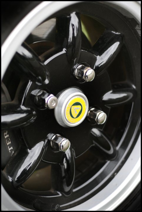 Back in April... - Page 1 - Caterham - PistonHeads - This is a close-up, high-resolution photograph of a tire hub cap featuring a central yellow circular logo with two lines of text around it, partially visible. The hub cap is part of a car's wheel assembly that appears to be coated in a glossy black material, which reflects some of the surrounding environment on its surface. The perspective of the camera angle is from above the hub cap, indicating the view is coming from above or looking over the top of the tire. The image captures the detail of the hub cap mechanically fastened to the wheel with visible bolts.