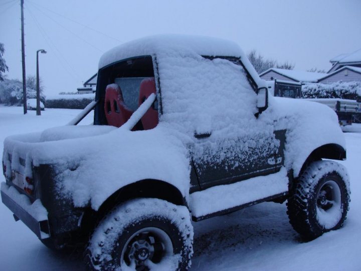 Snow - Page 101 - The Lounge - PistonHeads - The image features a pickup truck covered in a thick layer of snow, indicating recent or ongoing snowfall. The vehicle's windows are snow-covered, and red seats are visible through them. There are tire tracks in the surrounding snow, suggesting that the truck has been parked or driven on the snowy surface. The background, though dark, appears to be a static street scene with what might be other vehicles and possibly houses, but it is overshadowed by the dominance of the snow-covered truck.