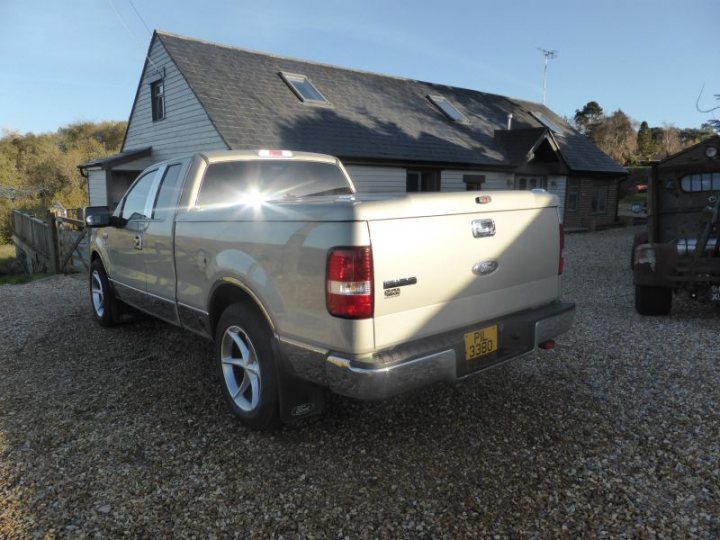 What To Look For When Buying Used 2007 F150? - Page 1 - Yank Motors - PistonHeads - This image features a silver Ford truck next to a wooden house with a grey shingle roof. The truck is parked on a gravel driveway. The sunlight shines brightly on the truck's rear, creating a contrast between the bright rear section and the shadowed front section. The license plate on the truck is clearly visible and it's daytime in the calm setting.