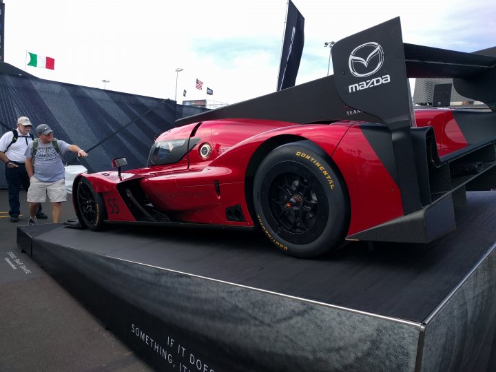 2018 Daytona 24hr - Page 4 - General Motorsport - PistonHeads - The image shows a stunning red Mazda endurance race car proudly displayed at a car show. The car, sleek and modern, is positioned prominently on a platform specifically designed for showcasing it. A few stats about the vehicle, such as its top speed and engine output, have been inscribed in white chalk on the platform, inviting spectators to learn more about this racing machine. In the background, spectators can be seen walking behind a typical concert-style infrastructure with black roofing and raised areas, adding to the ambiance of the event.