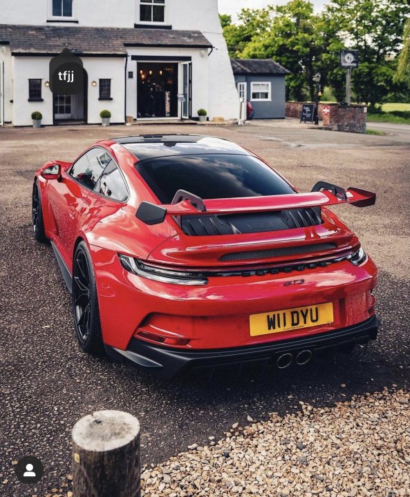 992 GT3 - Page 96 - 911/Carrera GT - PistonHeads UK - The image shows a red sports car parked in an outdoor area, possibly a driveway or parking lot. The car is positioned at an angle to the camera, emphasizing its sleek design and wide stance. It appears to be a high-performance vehicle, as suggested by its aggressive styling and large air intakes. There is a person visible in the background, partially obscured and dressed casually, suggesting they might be the owner or someone associated with the car. The environment includes a building with a white facade and a signboard with text that is too small to read clearly. The setting seems to be in a residential area, given the presence of a house and a driveway.