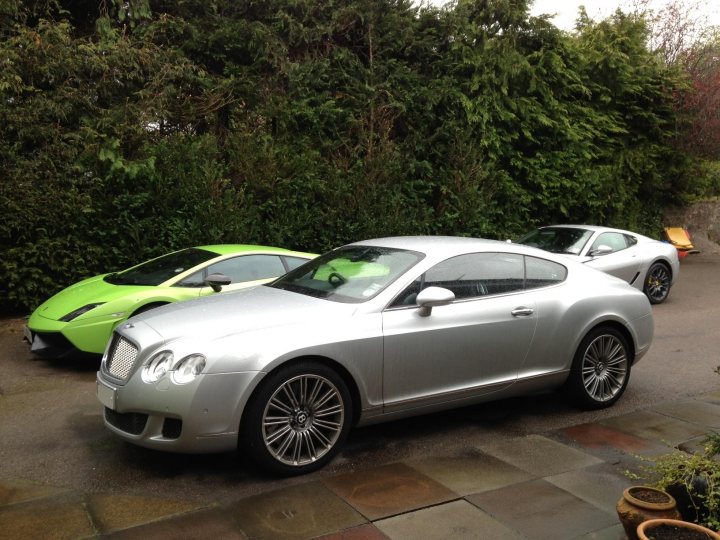 I’ve put my football boots on & bought a Bentley GT Speed - Page 1 - Rolls Royce & Bentley - PistonHeads - The image is a photograph showing two luxury cars parked next to each other on a paved surface. The car on the left is a bright green Bentley Flying Spur with a two-door design, while the car on the right is a silver Aston Martin with a sleek and aerodynamic appearance. The cars are positioned so that they are facing towards the left of the frame. They are parked on a driveway lined with neatly trimmed hedges. The driveway extends slightly into the background, providing a clear path to the parked vehicles. The image conveys a sense of luxury and exclusivity associated with the two car brands.