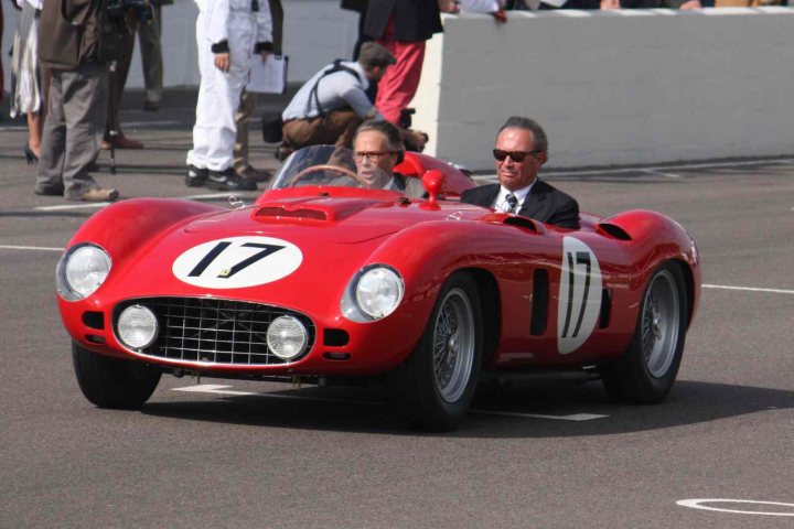 Revival images - Friday  - Page 1 - Goodwood Events - PistonHeads - The image depicts two men driving a small, vintage red sports car on a street. The driver, who is wearing glasses, appears focused on the road ahead. In the passenger seat, another man engages with the driver, contributing to the friendly atmosphere of the scene. To the side of the car, several individuals are observing and filming the ride. The backdrop of the image suggests an urban setting, with a low wall visible in the background.